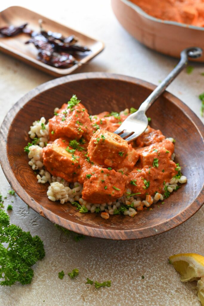 A fork piercing a piece of tofu in arrabiata sauce, with serrano peppers in the top of the image and parsley sprinkled over the top of the food.