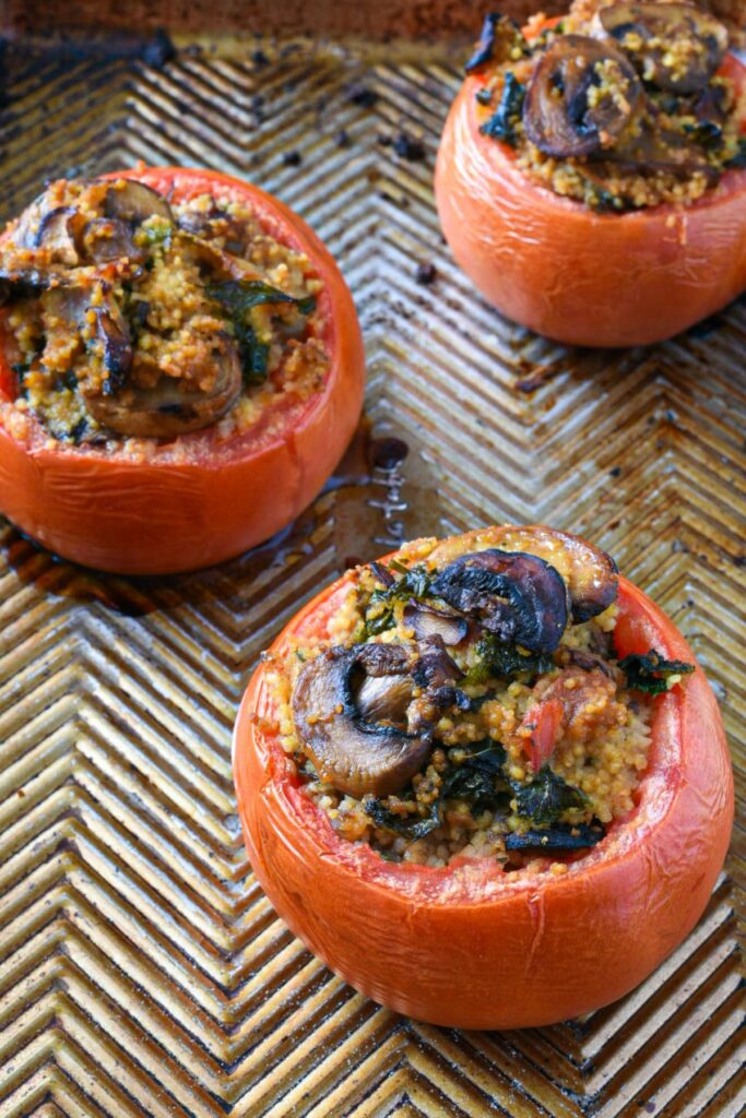 A close up image ofThree baked stuffed tomatoes on a ribbed baking sheet filled with mushrooms, couscous and kale mixture