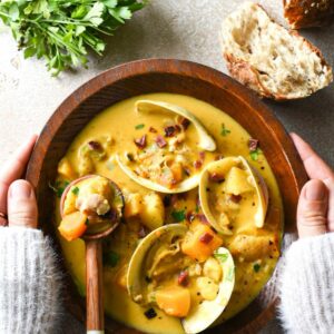 A wooden bowl of pumpkin clam chowder being held by two hands wearing a white sweater, with bread and parsley at the top of the image and clam shells in the soup.