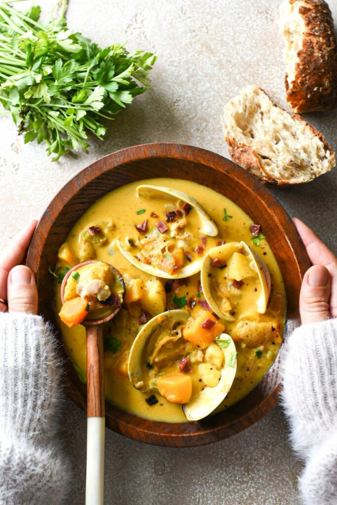 A wooden bowl of pumpkin clam chowder being held by two hands wearing a white sweater, with bread and parsley at the top of the image and clam shells in the soup.