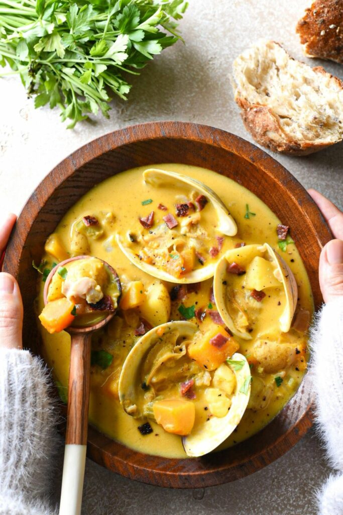 A close shot of a wooden bowl of pumpkin clam chowder being held by two hands wearing a white sweater, with bread and parsley at the top of the image and clam shells in the soup.