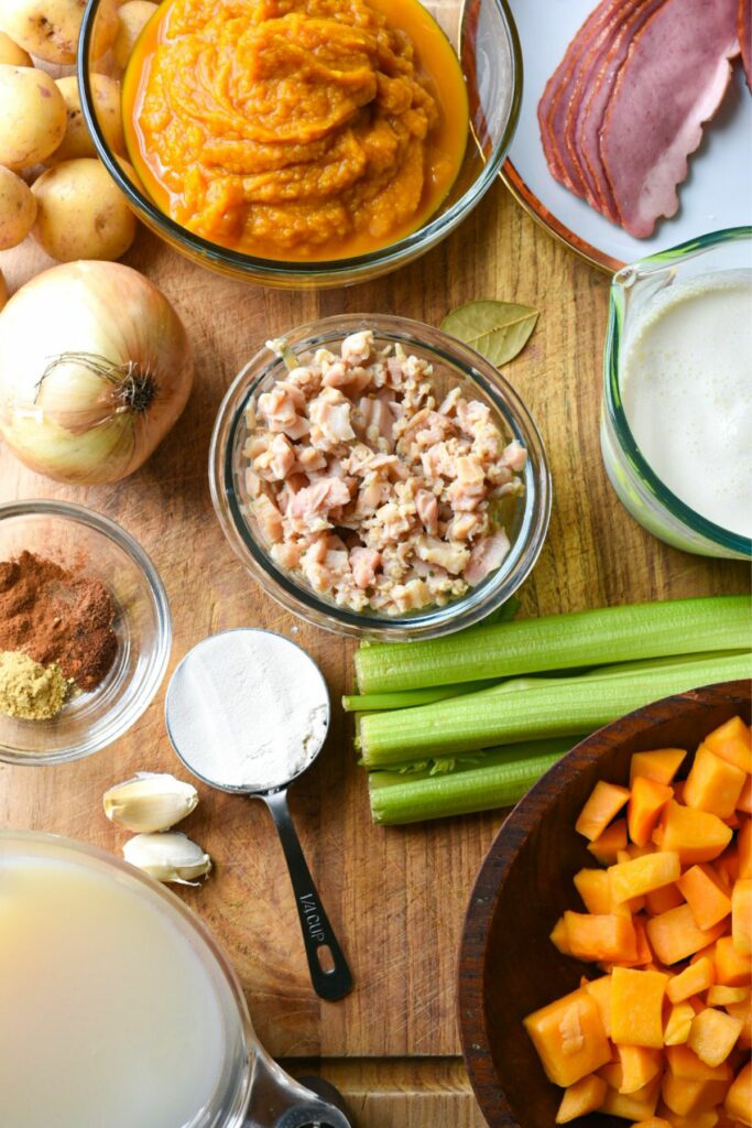 A wooden cutting board filled with ingredients for a fall soup recipe, including pumpkin puree, butternut squash, clams, bacon, onion, celery and cream.