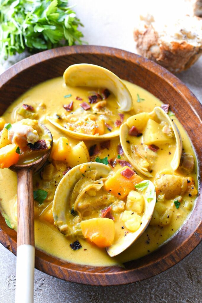 A wooden bowl filled with three open clam shells in a yellow pumpkin soup broth, with a wooden spoon filled with clams and potatoes.