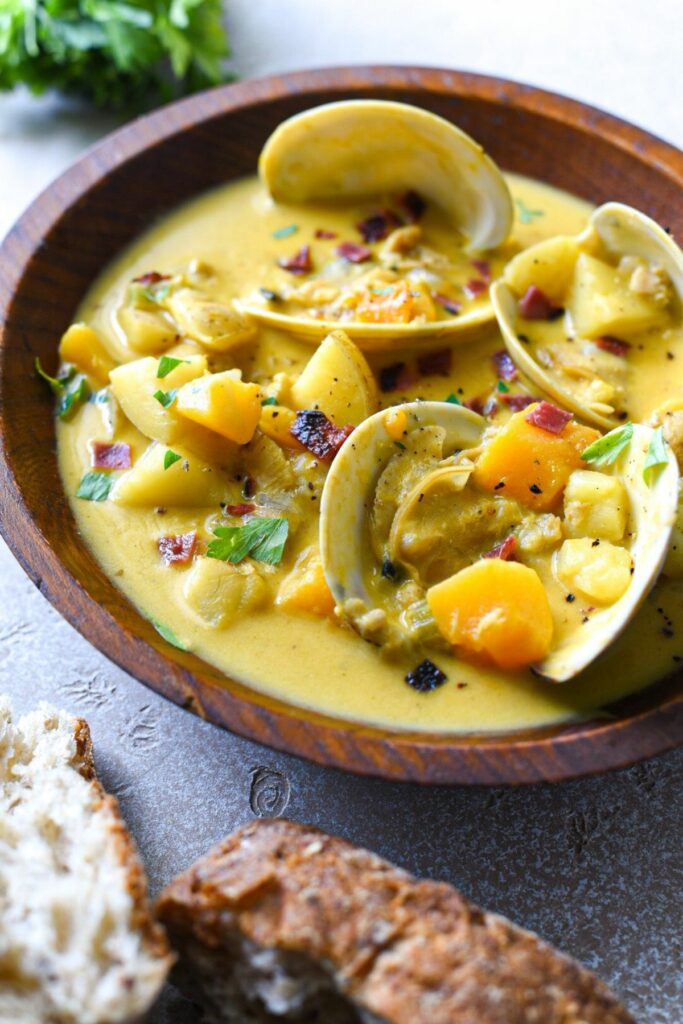 A wooden bowl filled with three clam shells in a yellow broth filled with potatoes, butternut squash, bacon and parsley.