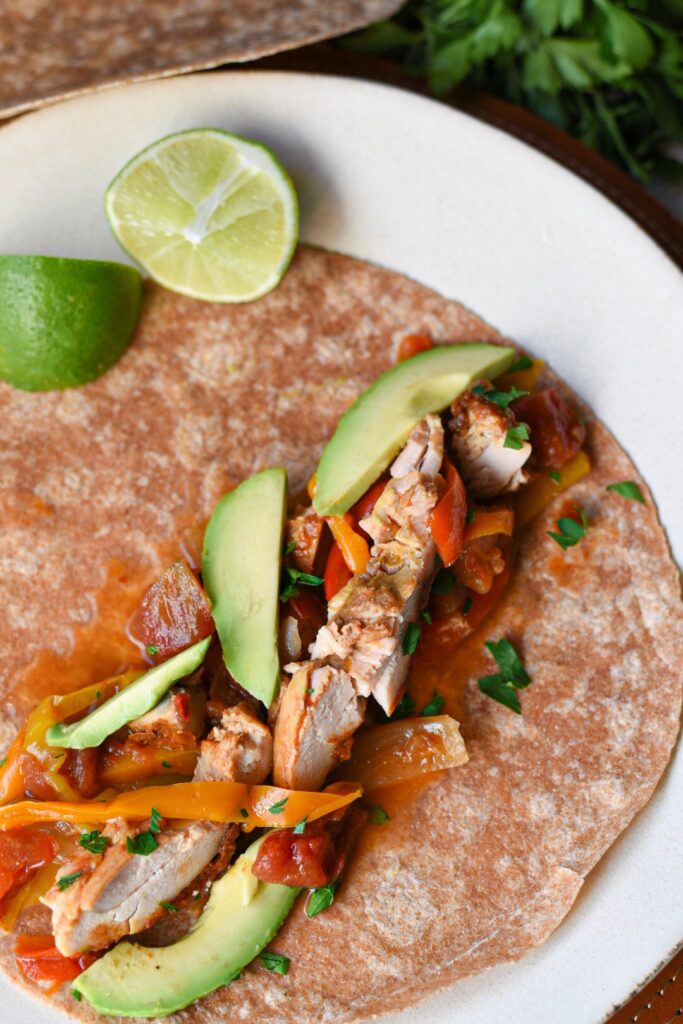 A whole wheat tortilla on a white plate filled with slow cooker chicken fajitas topped with sliced avocado