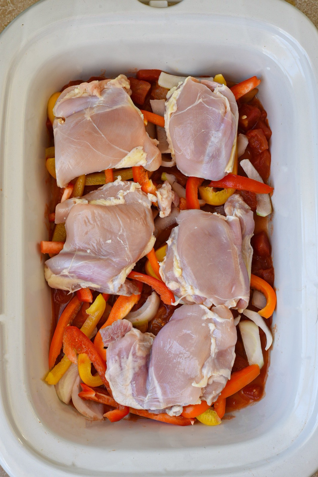 A white crockpot filled with colorful bell peppers, onion, tomato and topped with chicken thighs