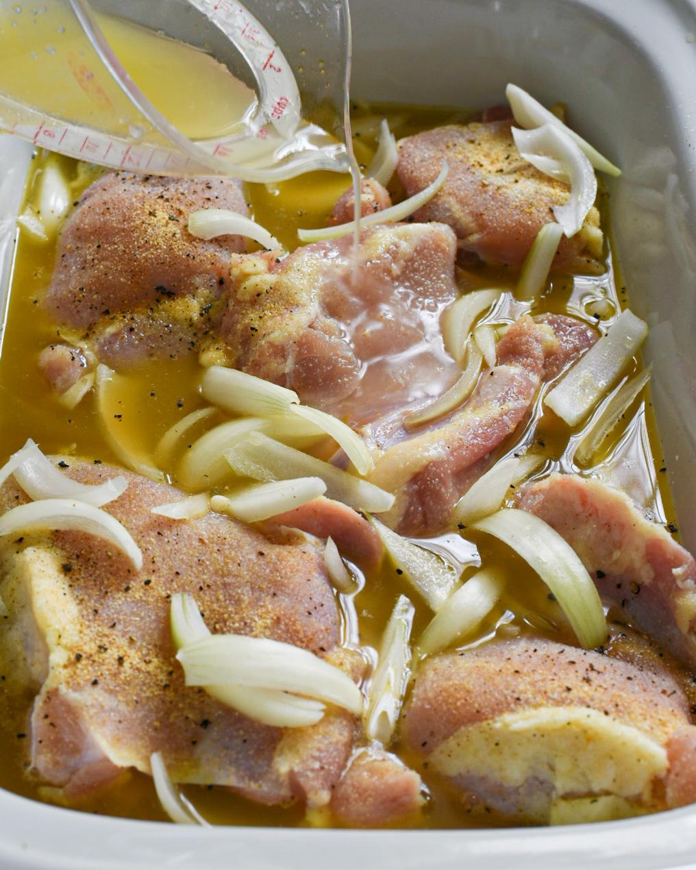 Raw chicken thighs in a slow cooker topped with sliced onion and chicken broth