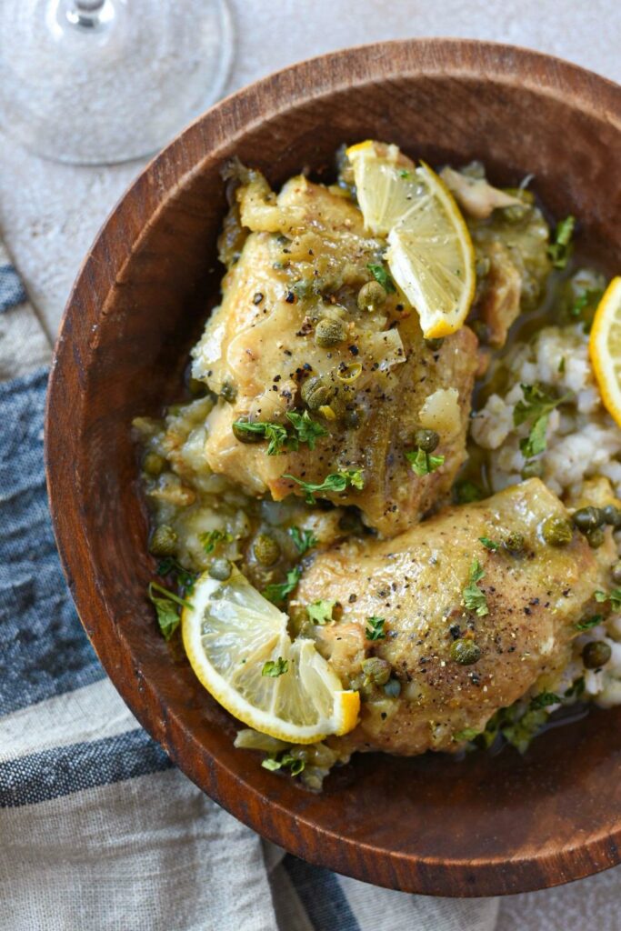 Half of a wooden bowl filled with barley and cooked chicken thighs topped with lemon slices, capers and pepper.