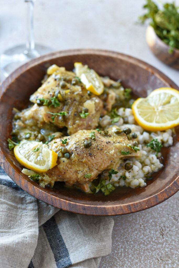A wooden bowl filled with barley and topped with crockpot lemon chicken thighs covered in capers