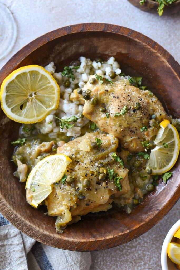 A wooden bowl filled with slow cooked chicken thighs with lemon slices and fresh cracked black pepper over the top. There is barley in the bowl