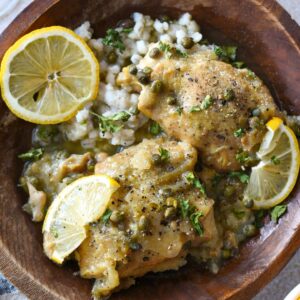 A wooden bowl filled with slow cooked chicken thighs with lemon slices and fresh cracked black pepper over the top. There is barley in the bowl