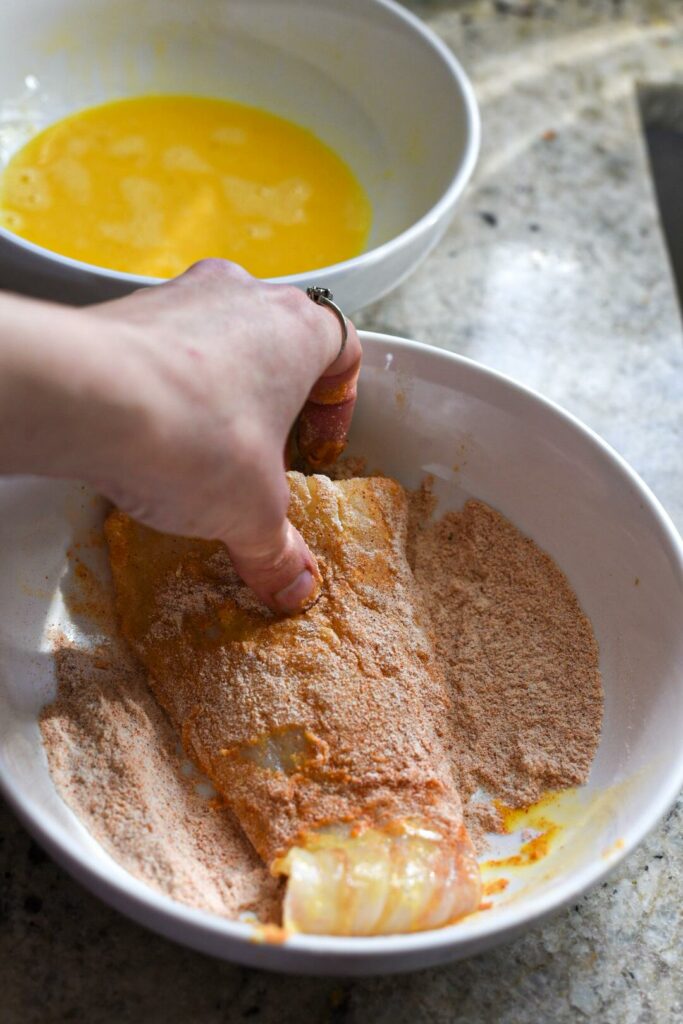 Scrod fish being dredged in a brown rice flour and spice mixture after being dipped in egg