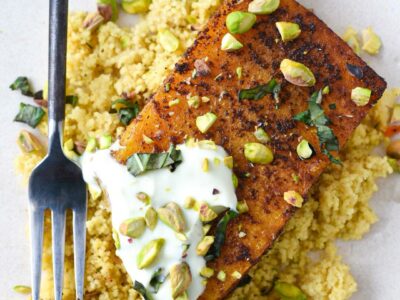 A roasted butternut squash steak with yogurt sauce, pistachios and basil over a bed of couscous and a fork showing a piece cut off