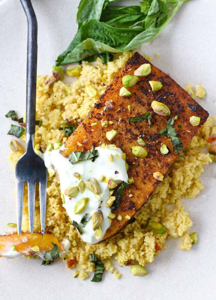 A roasted butternut squash steak with yogurt sauce, pistachios and basil over a bed of couscous and a fork showing a piece cut off