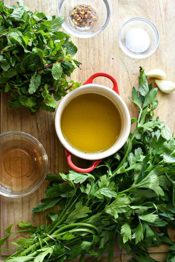A wooden cutting board with a small bowl of olive oil, salt, red pepper flakes, garlic, vinegar, parsley, and mint