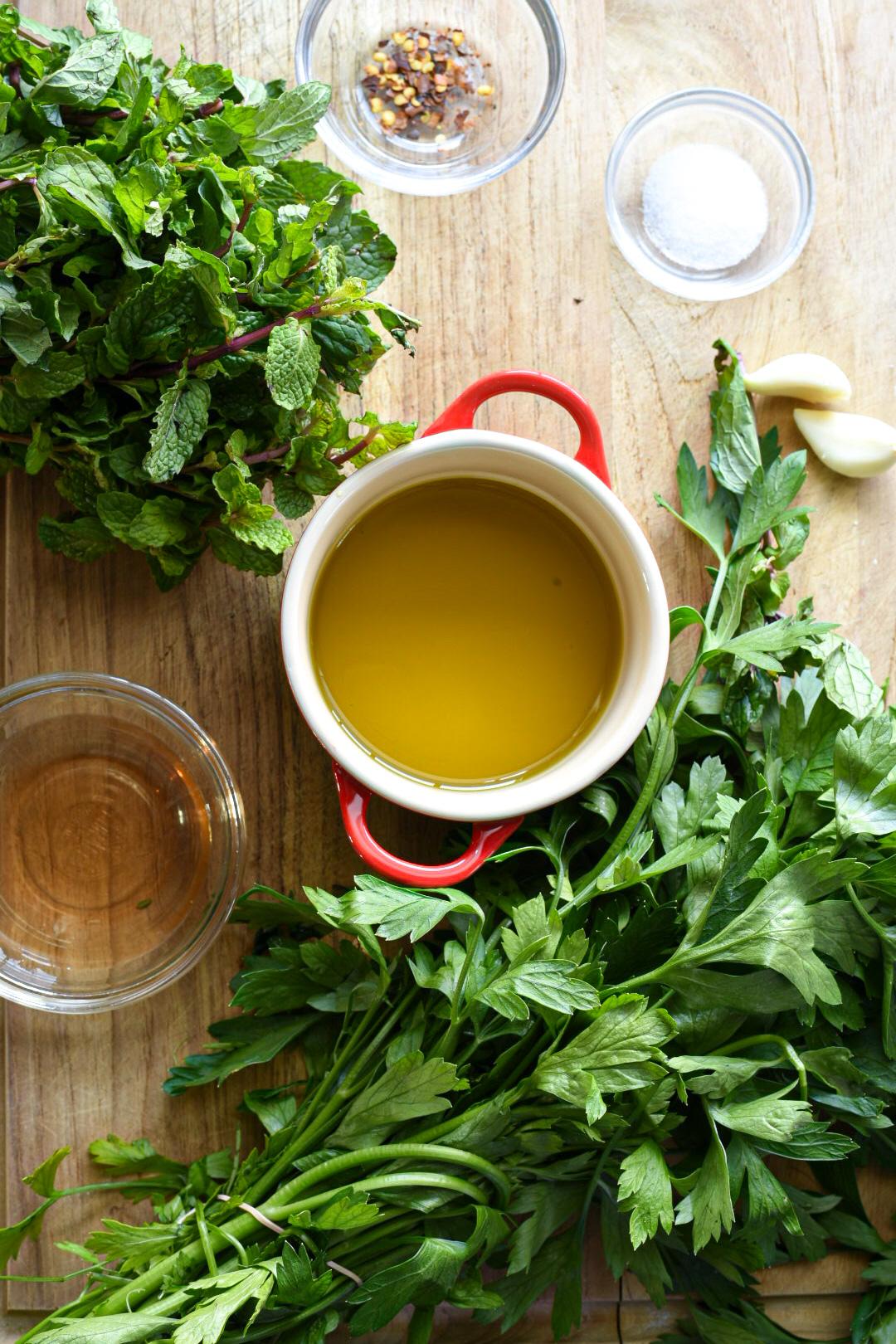 A wooden cutting board with a small bowl of olive oil, salt, red pepper flakes, garlic, vinegar, parsley, and mint