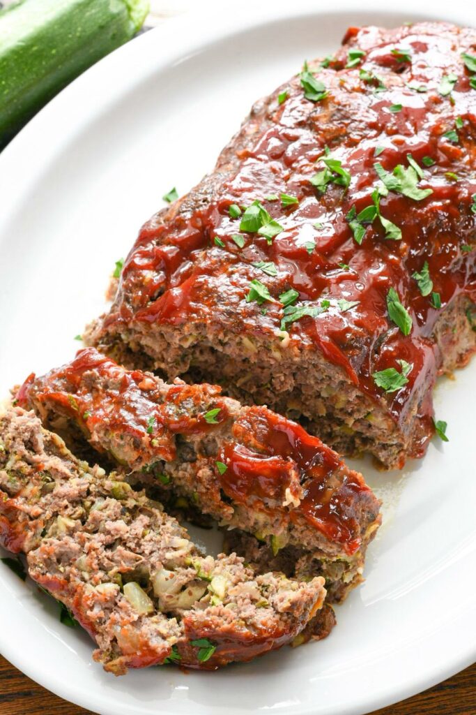 A finished zucchini meatloaf recipe with two slices and a log on a white serving platter sprinkled with parsley