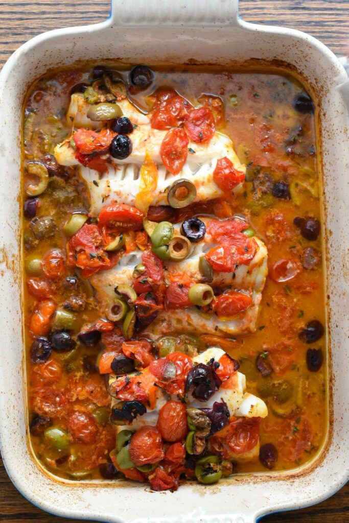 Overhead shot of three baked cod fillets in a baking dish covered in olives, tomatoes and a lemony sauce