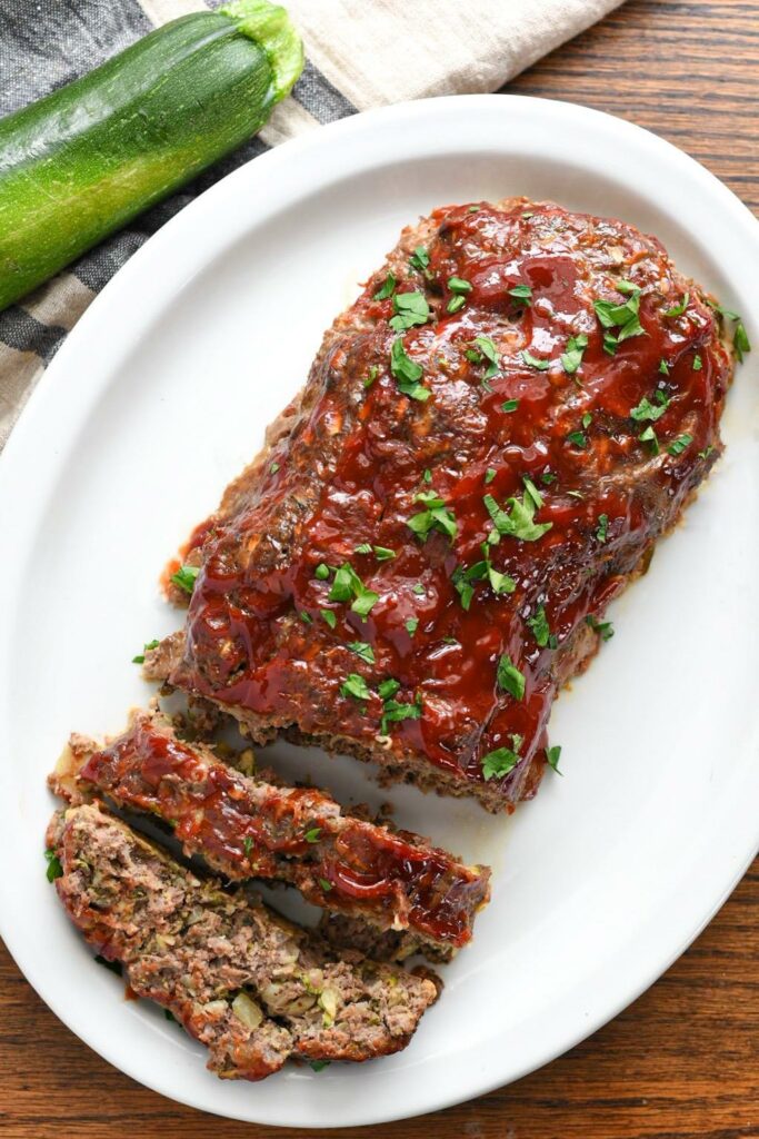 Overhead shot of a meatloaf with ketchup sauce on a white platter with a zucchini in the corner