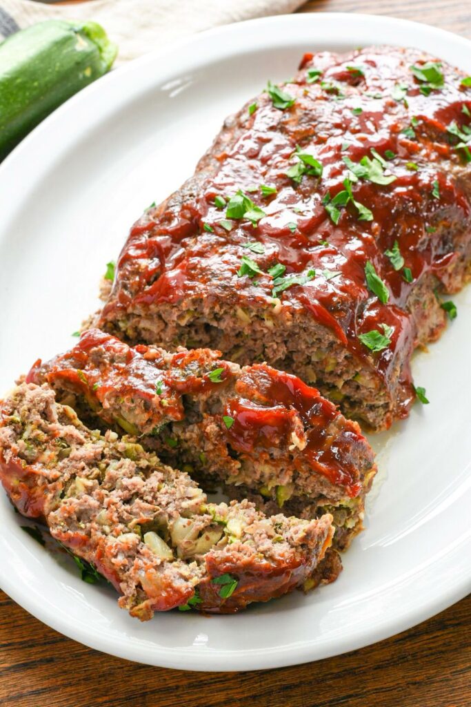 Sliced meatloaf on a white serving platter with pieces of zucchini visible and a bright red sauce