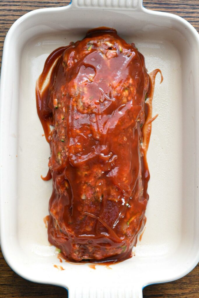 A freshly sauced raw meatloaf in a white baking dish with bright red ketchup sauce over the top