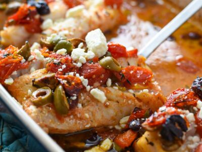 A spatula taking a piece of mediterranean baked cod topped with tomatoes, olives and feta out of a blue baking dish