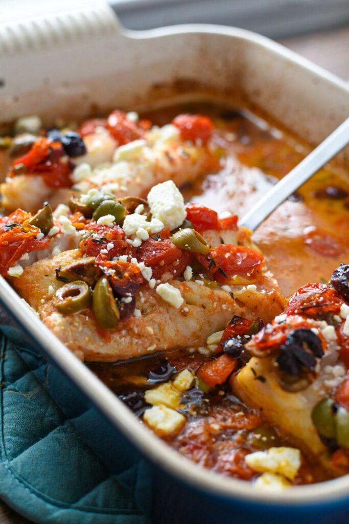 A spatula taking a piece of mediterranean baked cod topped with tomatoes, olives and feta out of a blue baking dish