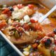 A spatula taking a piece of mediterranean baked cod topped with tomatoes, olives and feta out of a blue baking dish