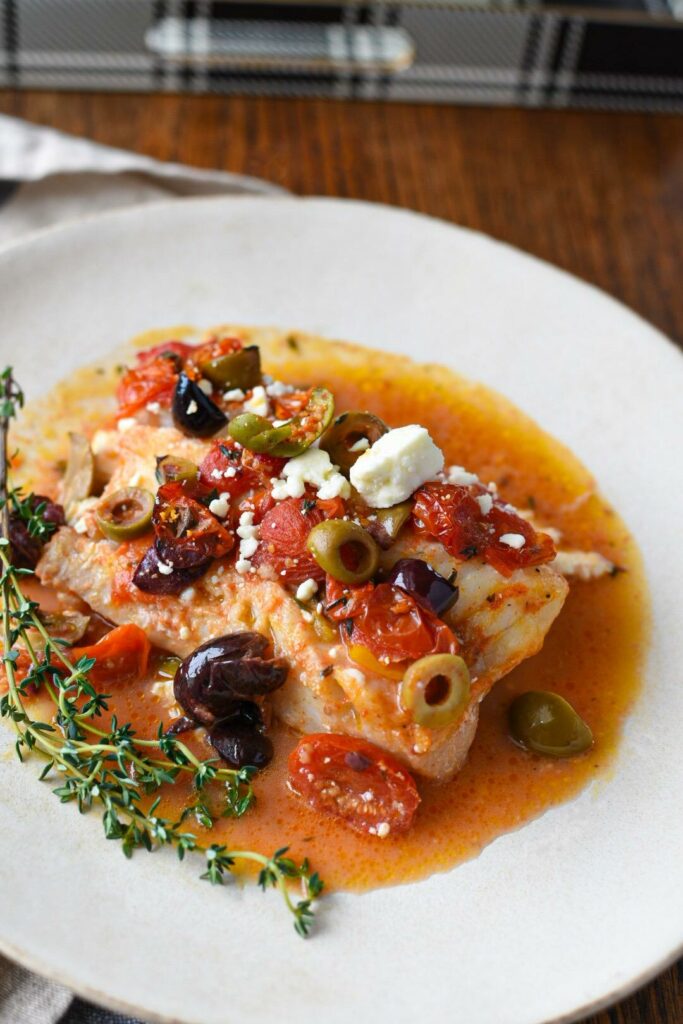 Mediterranean baked cod recipe on a white plate topped with green and black olives, tomatoes and feta with a sprig of thyme