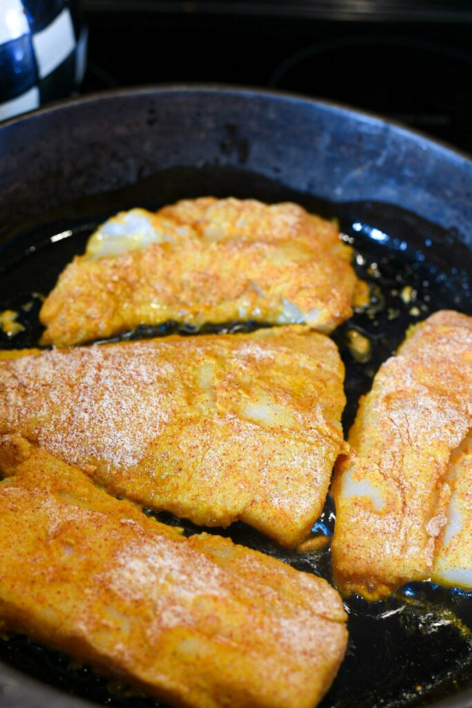 Scrod being pan seared in a black cast iron skillet after dredged in egg and spices