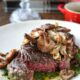 A cooked sous vide sirloin steak cut into to see the medium rare pink inside, topped with wild mushrooms and chimichurri sauce