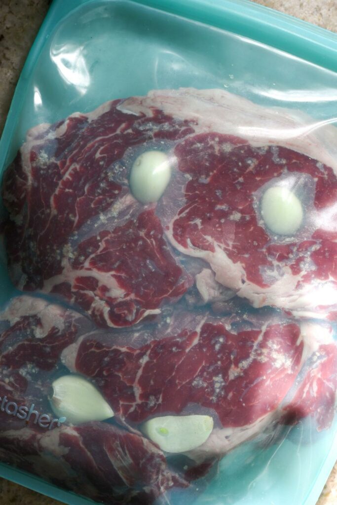 A blue stasher bag filled with raw sirloin steak and garlic cloves