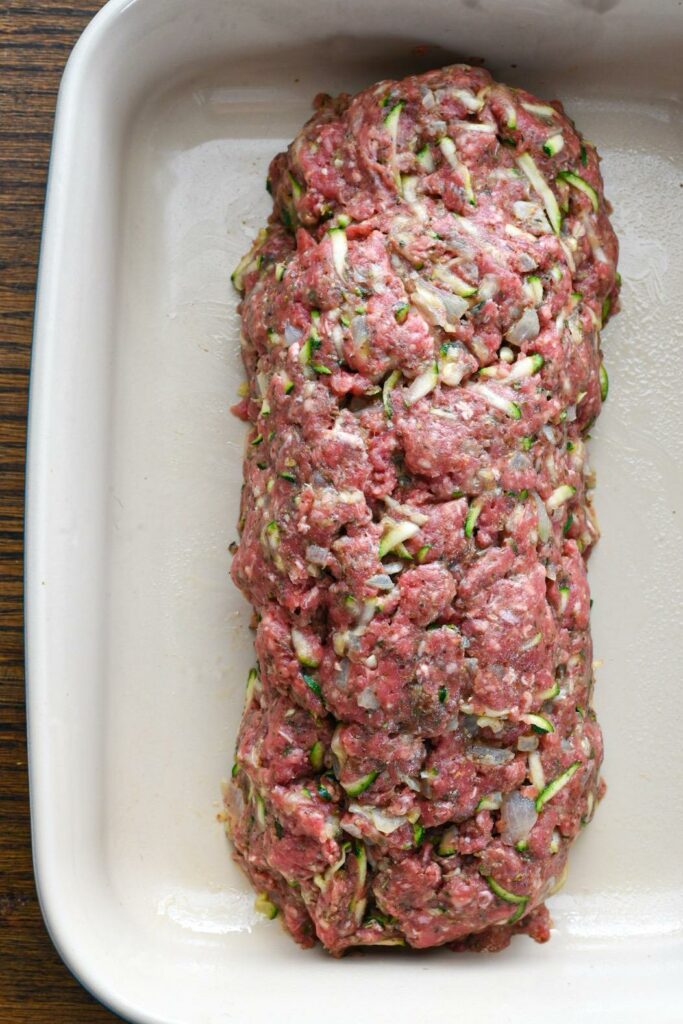A raw loaf of meatloaf in a baking dish with shredded zucchini visible in the loaf