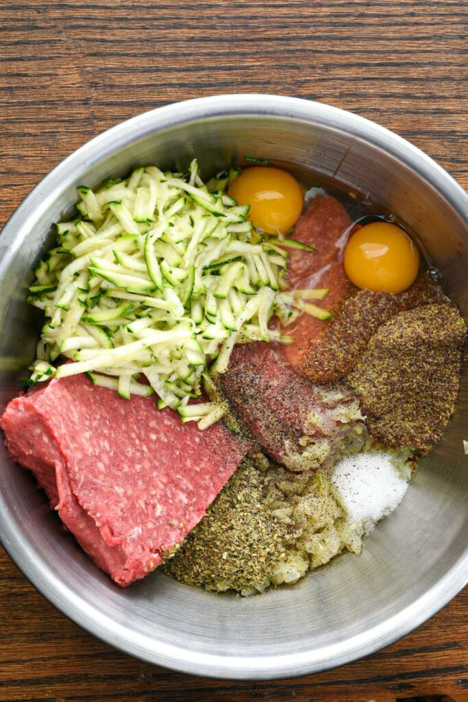 A mixing bowl filled with raw beef, shredded zucchini, eggs, flax meal, spices and onions