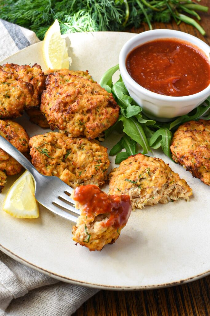 A bite of air fryer crab cake on a fork with cocktail sauce on it and more crab cakes on a plate in the background