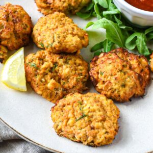 Seven golden crab cakes on a white plate with arugula, a lemon slice and cocktail sauce