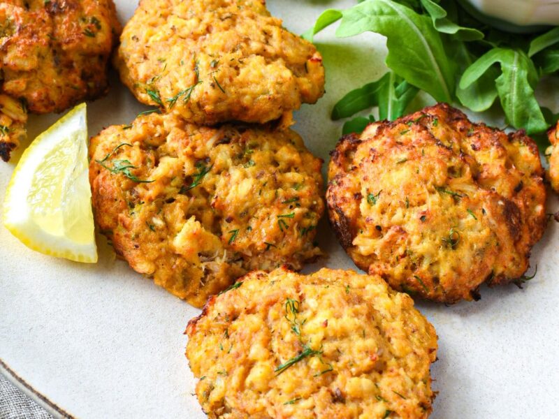 Seven golden crab cakes on a white plate with arugula, a lemon slice and cocktail sauce
