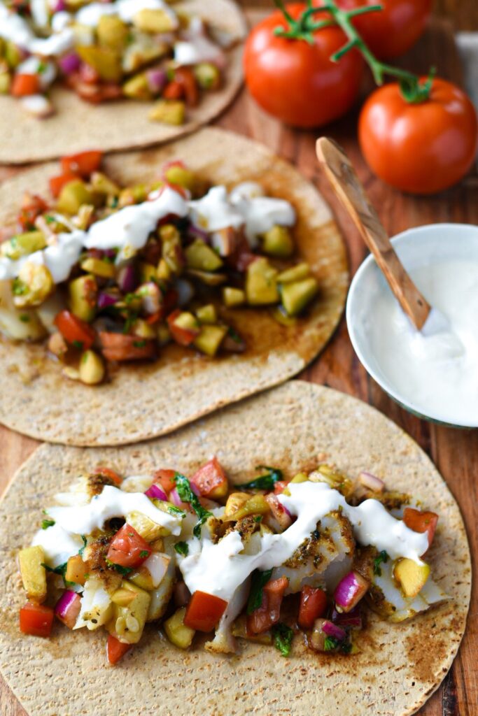 Three Mediterranean baked fish tacos on a wooden board topped with cucumber tomato balsamic salad and a lemon garlic yogurt sauce