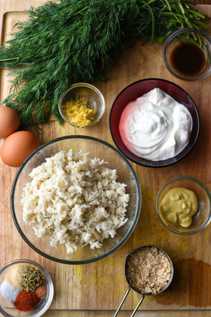 The ingredients for crab cakes in bowls on a wooden cutting board with a bunch of dill at the top
