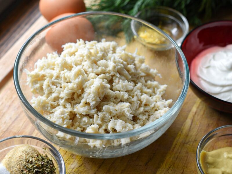 A wooden cutting board with a glass bowl filled with crab meat surrounded by more ingredients to make crab cakes