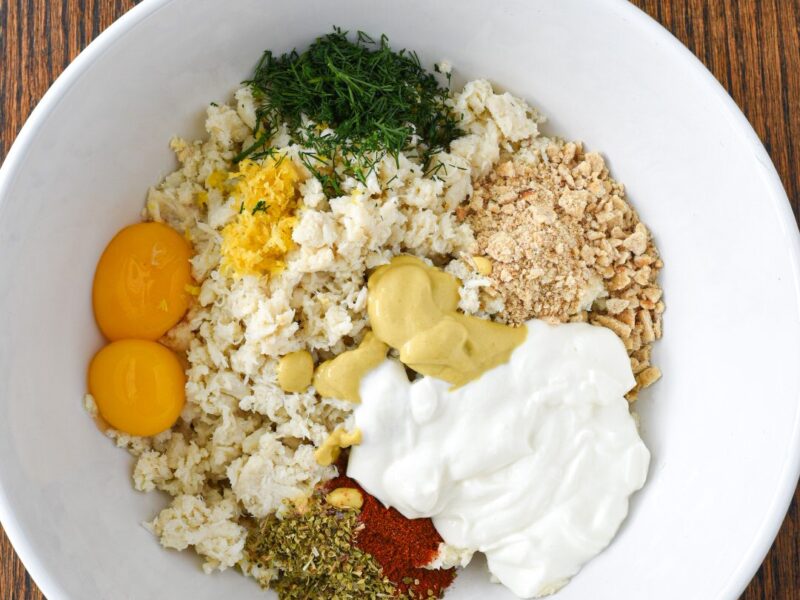 A large white bowl filled with the ingredients to make crab cakes prior to being mixed, including crab, dill, crackers, yogurt, mustard, spices and egg yolks