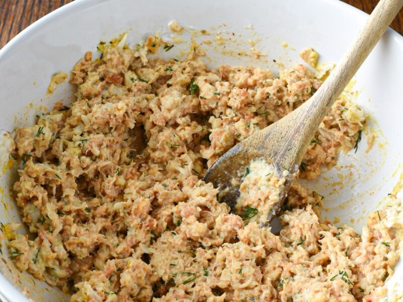 A bowl of a crab cake recipe mixed together with a wooden spoon with a bunch of dill in the background