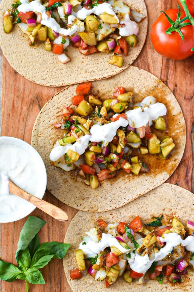 Three in focus Mediterranean baked fish tacos on a wooden board topped with cucumber tomato balsamic salad and a lemon garlic yogurt sauce