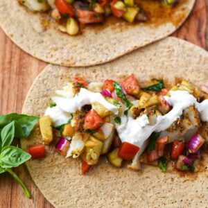 Two large tortillas on a wooden board topped with baked fish, cucumber tomato balsamic salad and yogurt sauce