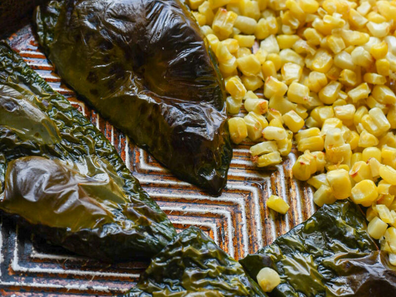 A close up image of blistered poblano peppers and browned corn on a brown baking sheet.