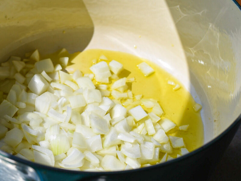 A large dutch oven in the sunlight filled with olive oil and chopped onions