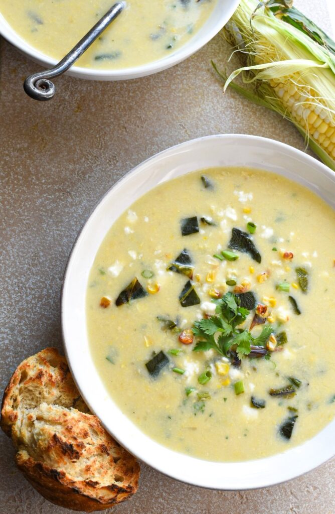 An overhead shot of a white bowl filled with a poblano corn chowder recipe with another bowl above it and a piece of crusty bread next to the bowl