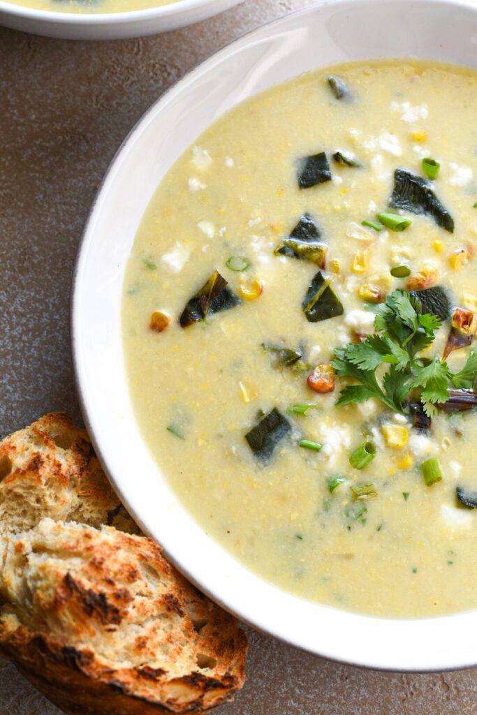 A white bowl filled with poblano corn chowder with feta, cilantro and green onion on top and a piece of crusty bread on the side