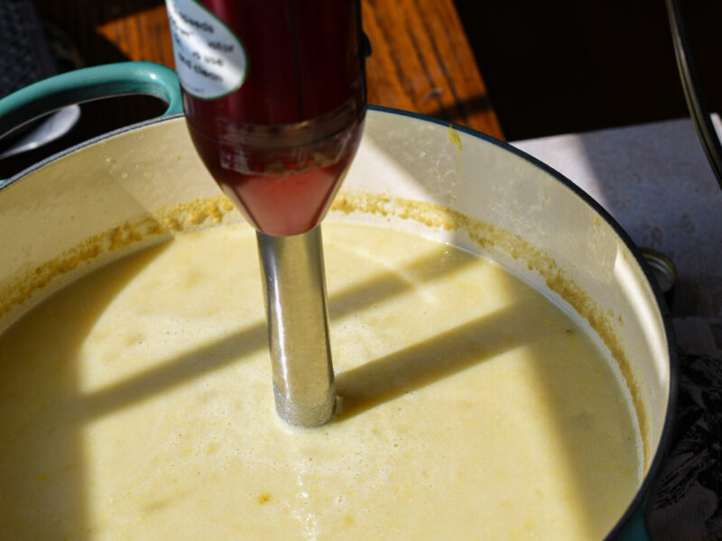A large blue dutch oven with poblano corn chowder in it being blended by a hand blender