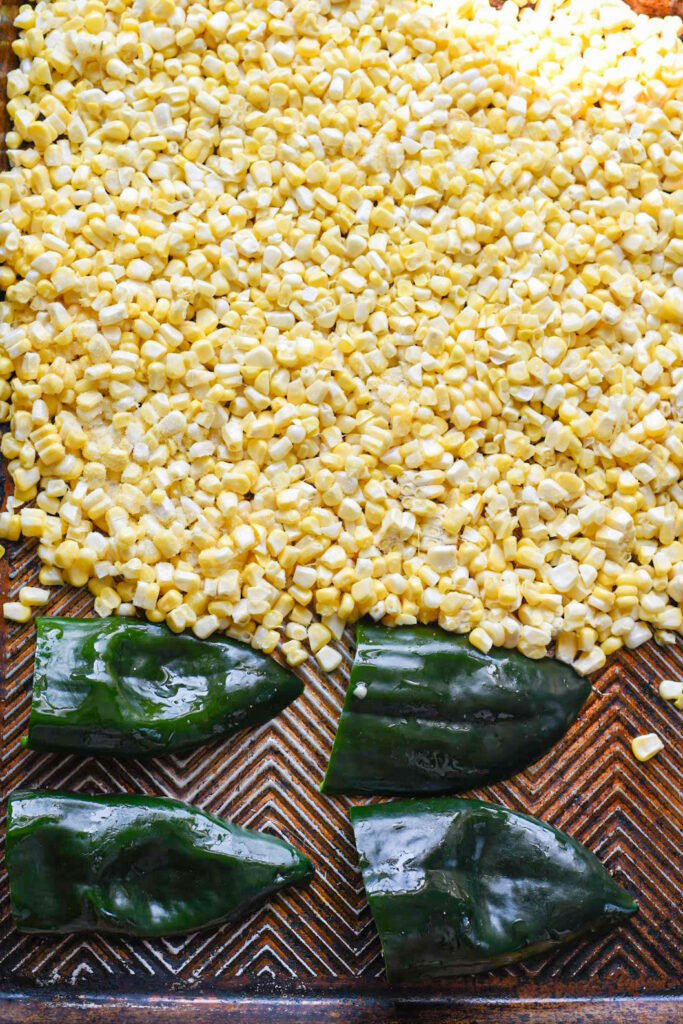 Fresh corn kernels spread out on a metal baking dish with 4 poblano pepper halves on the baking dish